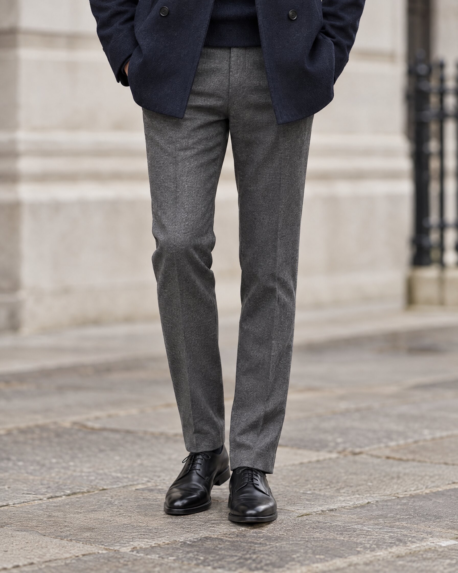 Man in slim shark grey wool trousers and a navy peacoat, cropped at the waist, on a city street