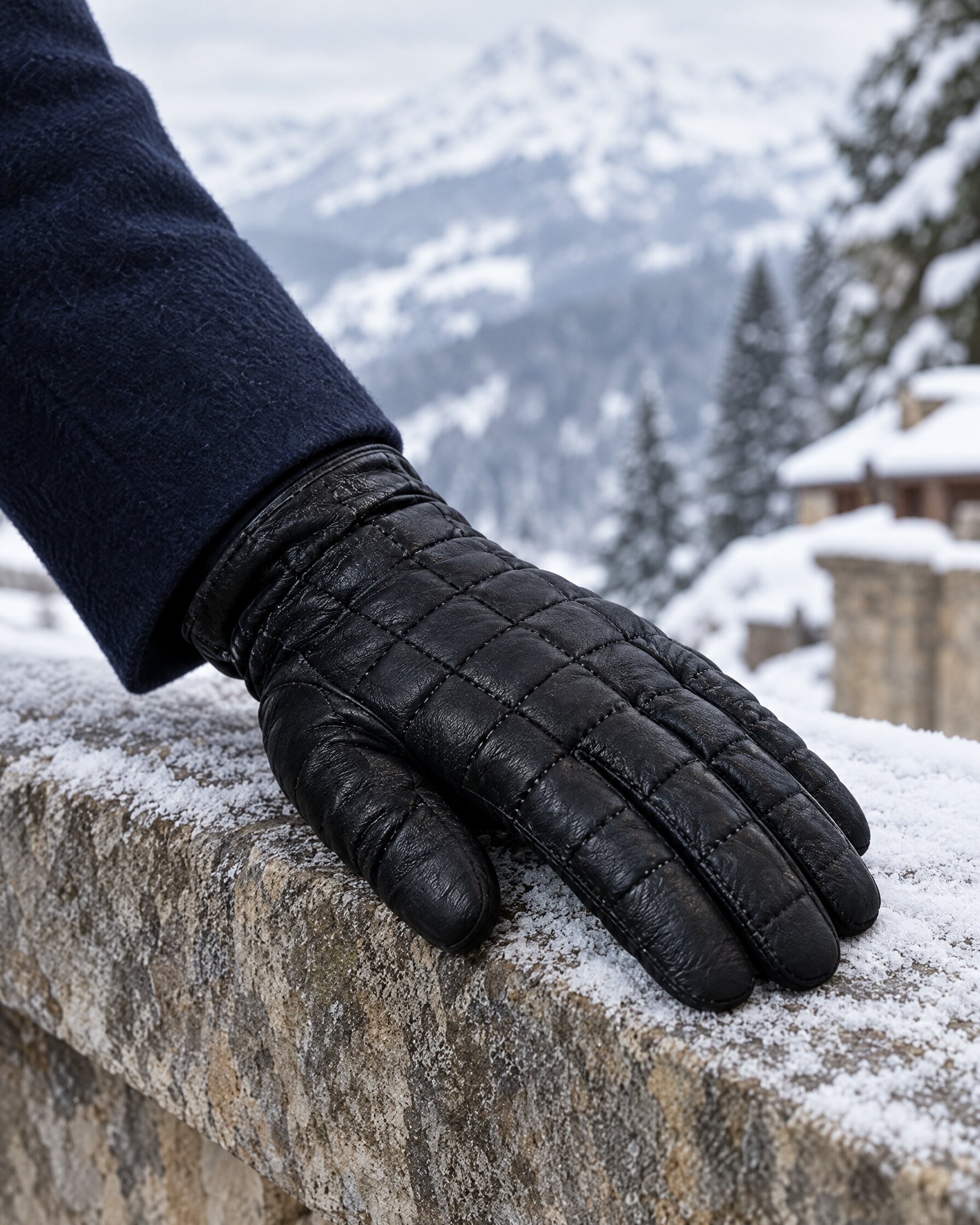 Hand in a black quilted lambskin glove resting on a stone wall in the snow, navy overcoat cuff visible