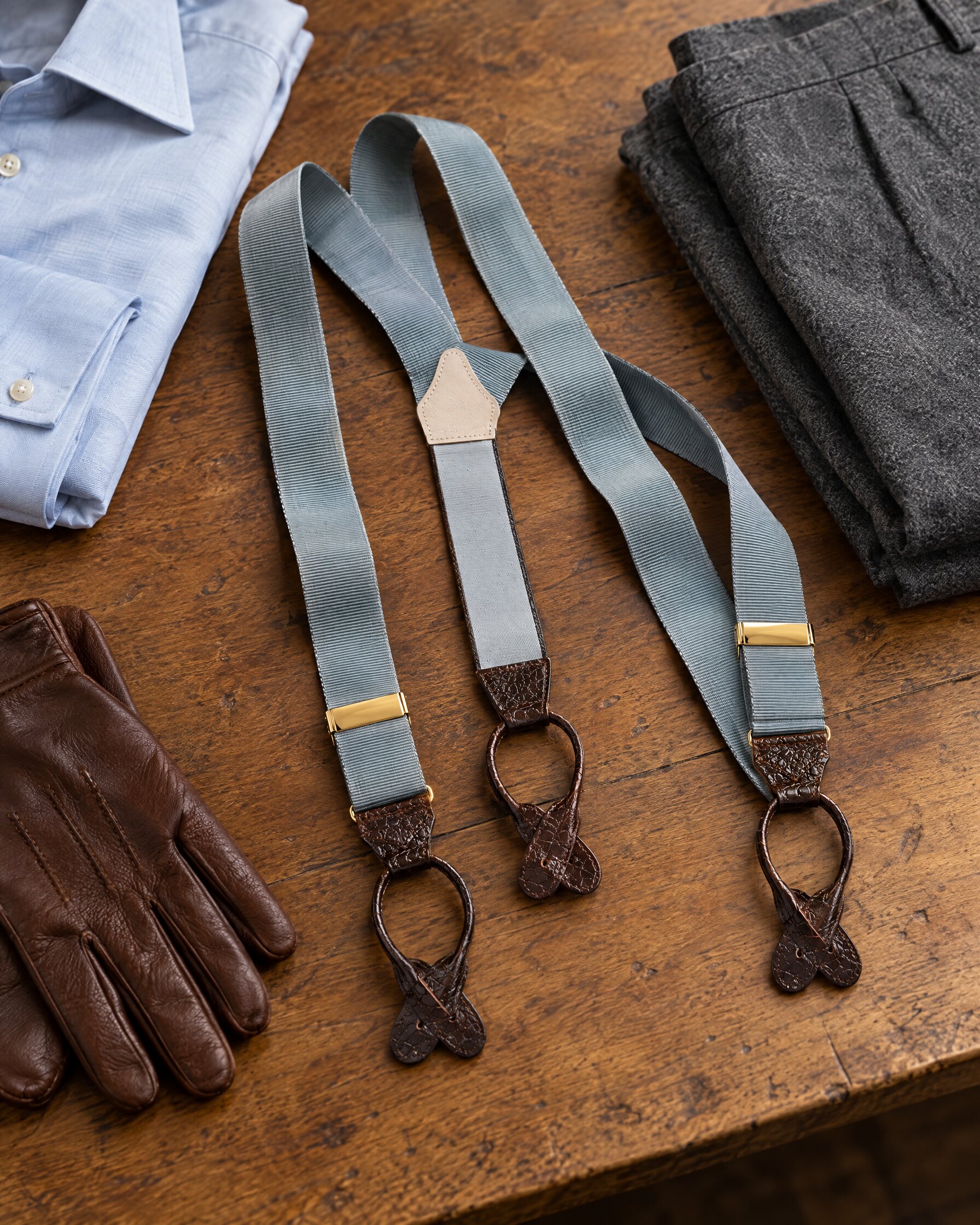 Albert Thurston dove grey braces with brown leather ends and gilt clasps laid flat beside a pale blue shirt