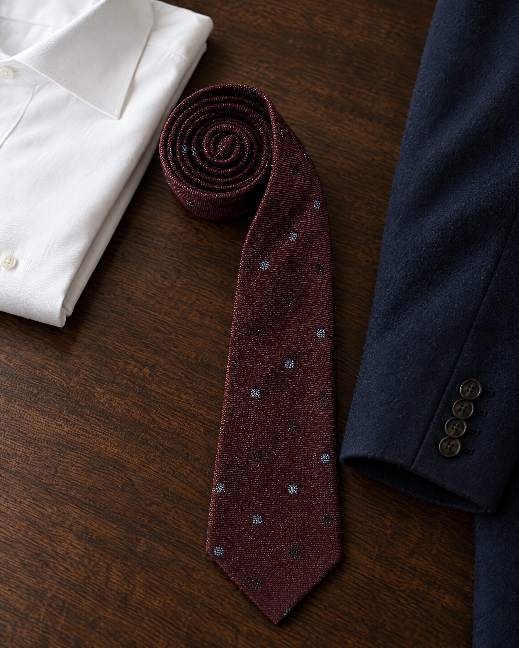 Burgundy silk twill Alexander Olch Avery necktie with bourette spots laid on dark oak beside a white shirt