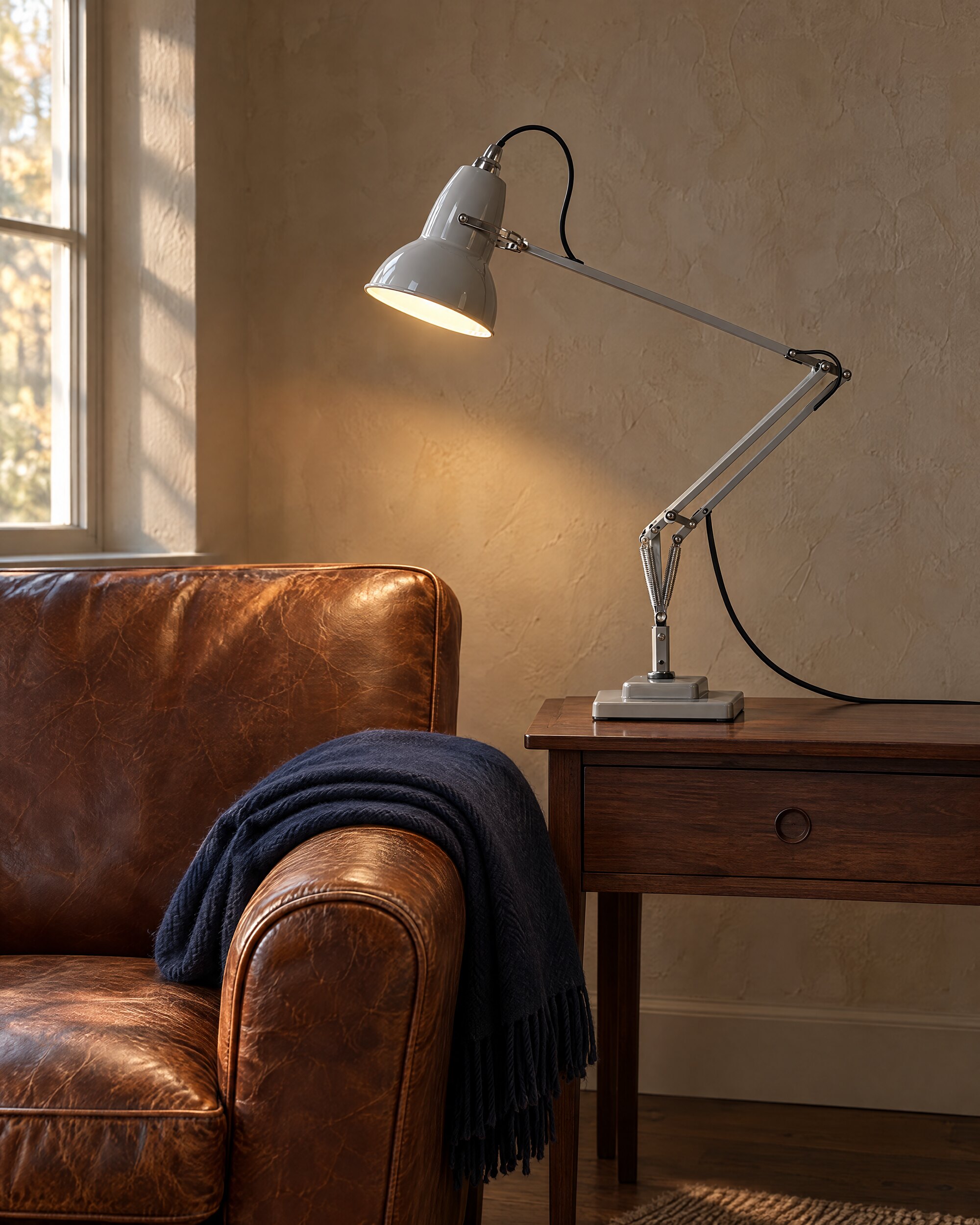 Pale grey Anglepoise desk lamp beside a leather armchair in a quiet, sparsely furnished room