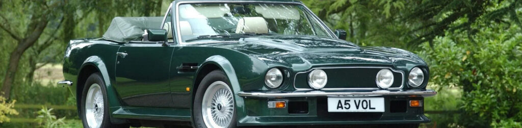 Close-up of the front of a classic Aston Martin V8 Vantage in dark grey, showing the long bonnet and deep front spoiler