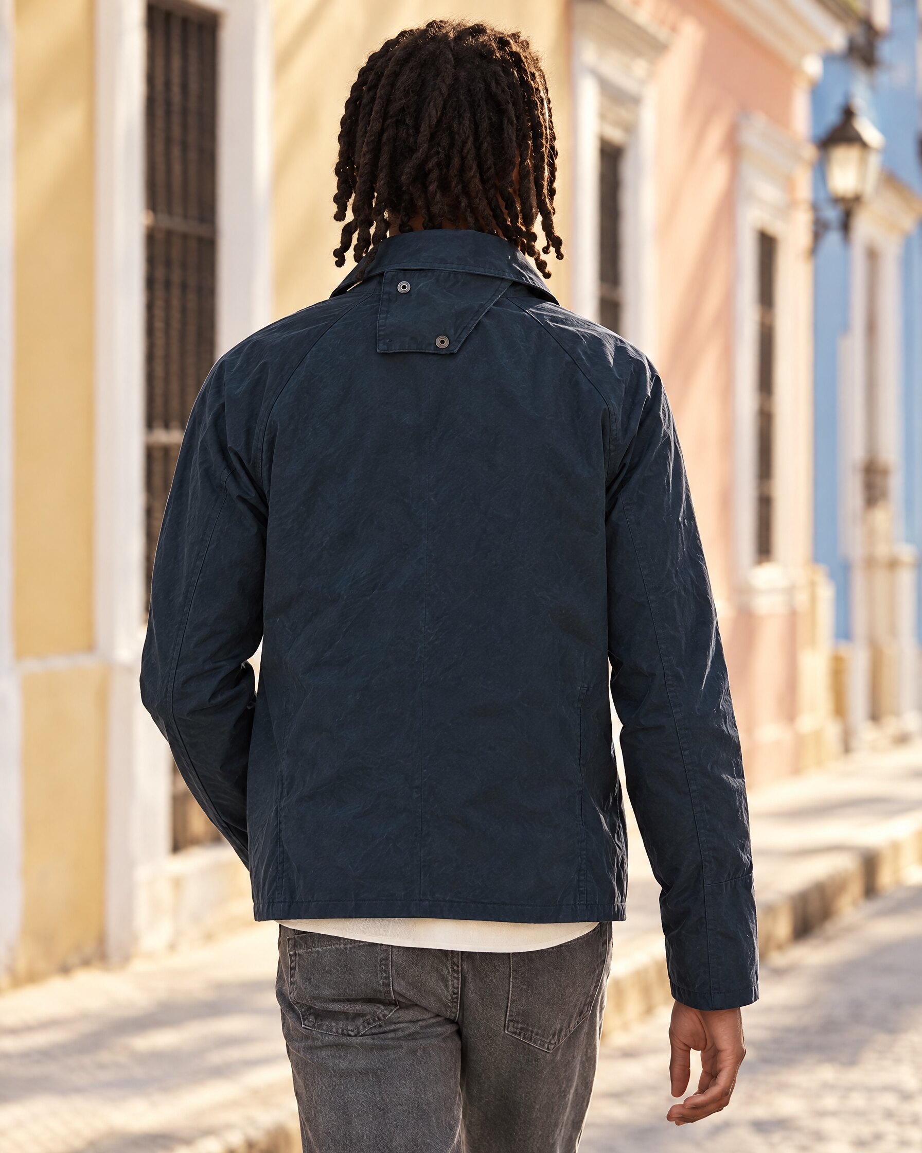 Man in a navy waxed cotton short jacket walking along a sunlit street, seen from behind