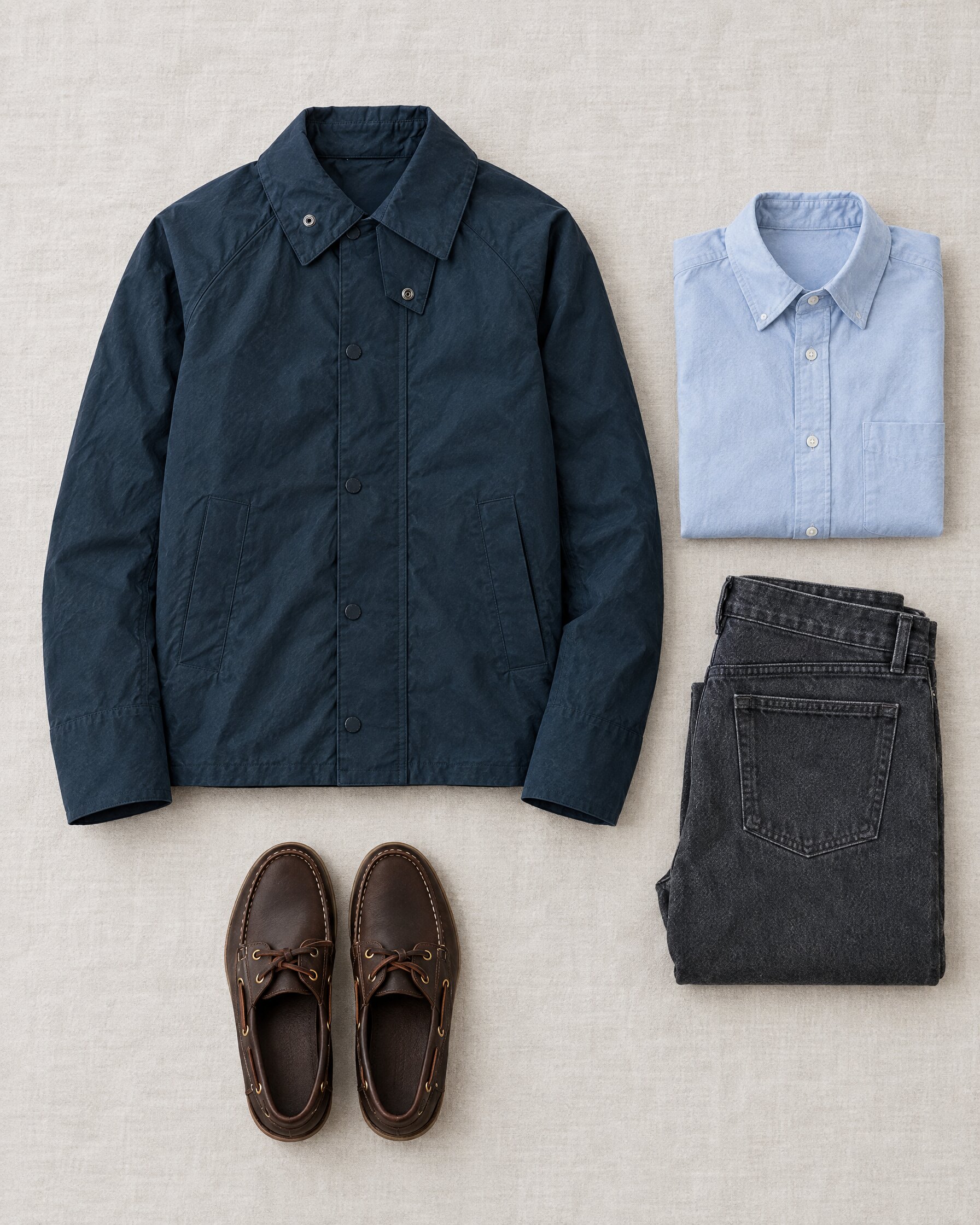 Navy waxed cotton Barbour Graham jacket laid flat with a pale blue shirt, grey jeans and brown boat shoes