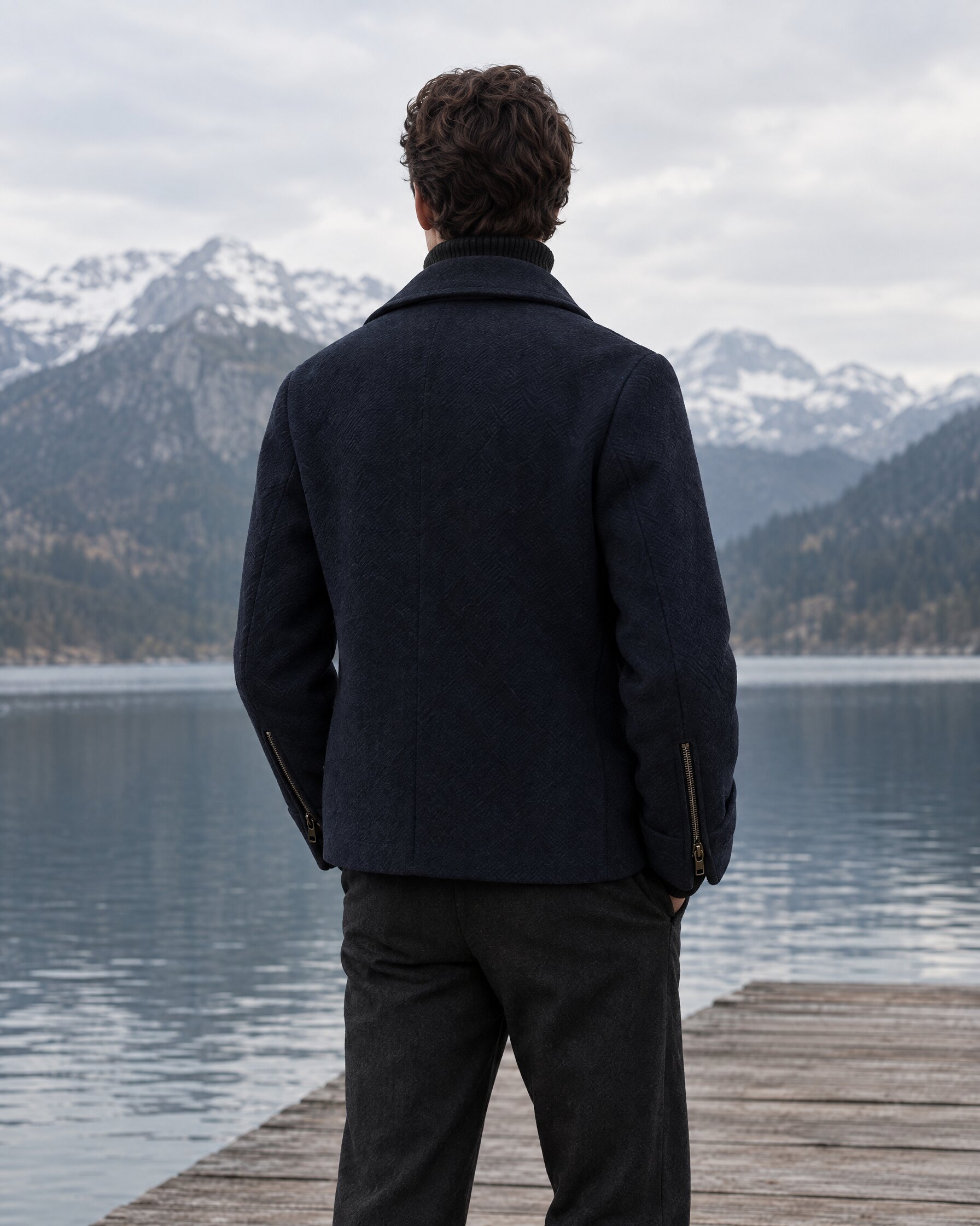 Man in a navy Dior Homme Spectre jacket-style peacoat with zippered sleeves on a lakeside jetty, seen from behind