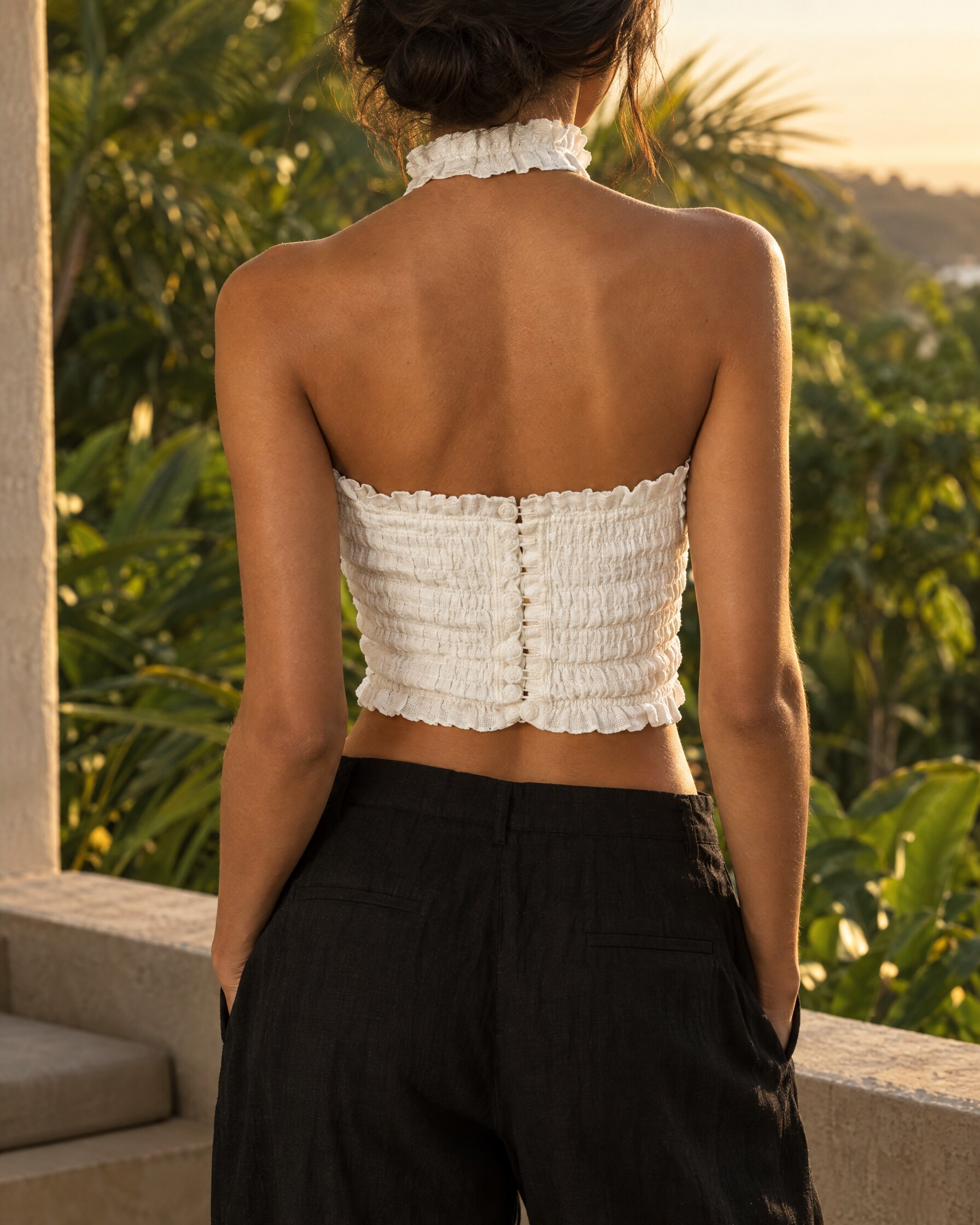 White linen halter neck top with ruffled collar and hook and eye back fastening, worn with black trousers on a shaded terrace