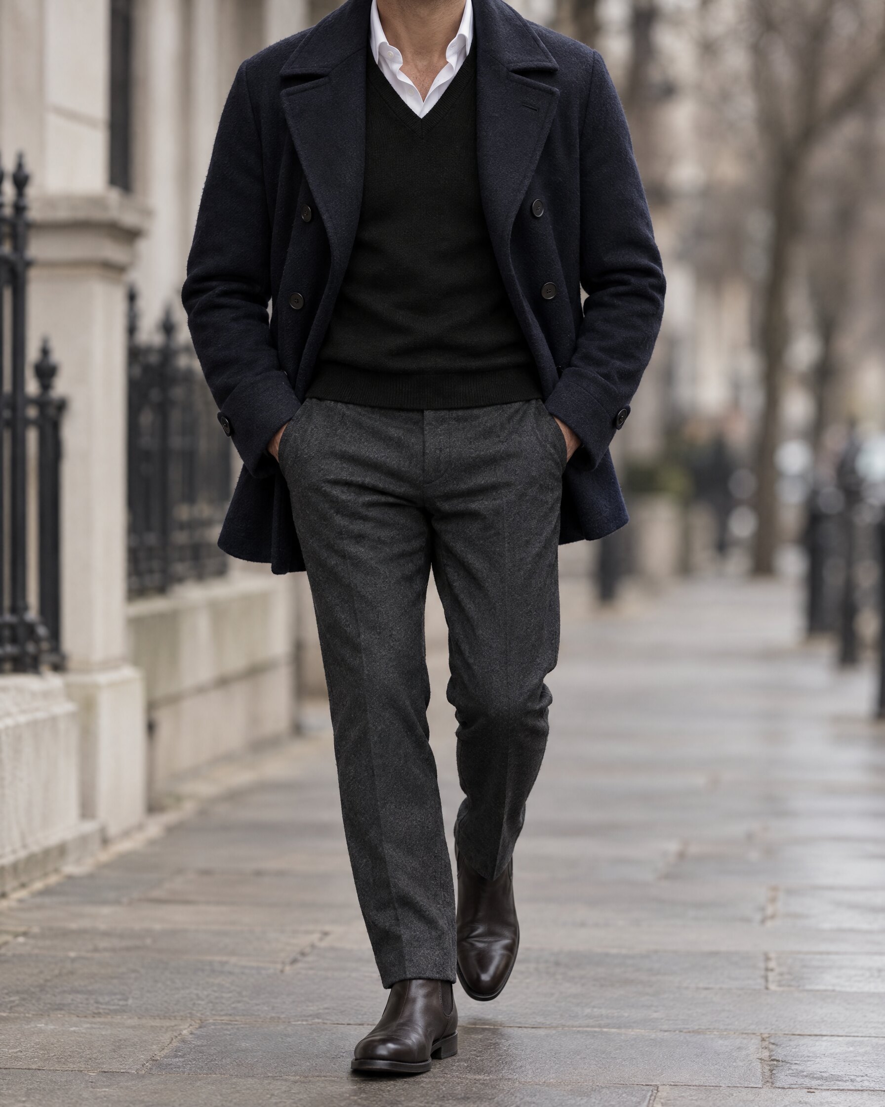 Man in a black merino v-neck jumper under an open navy peacoat on a city street, face out of frame