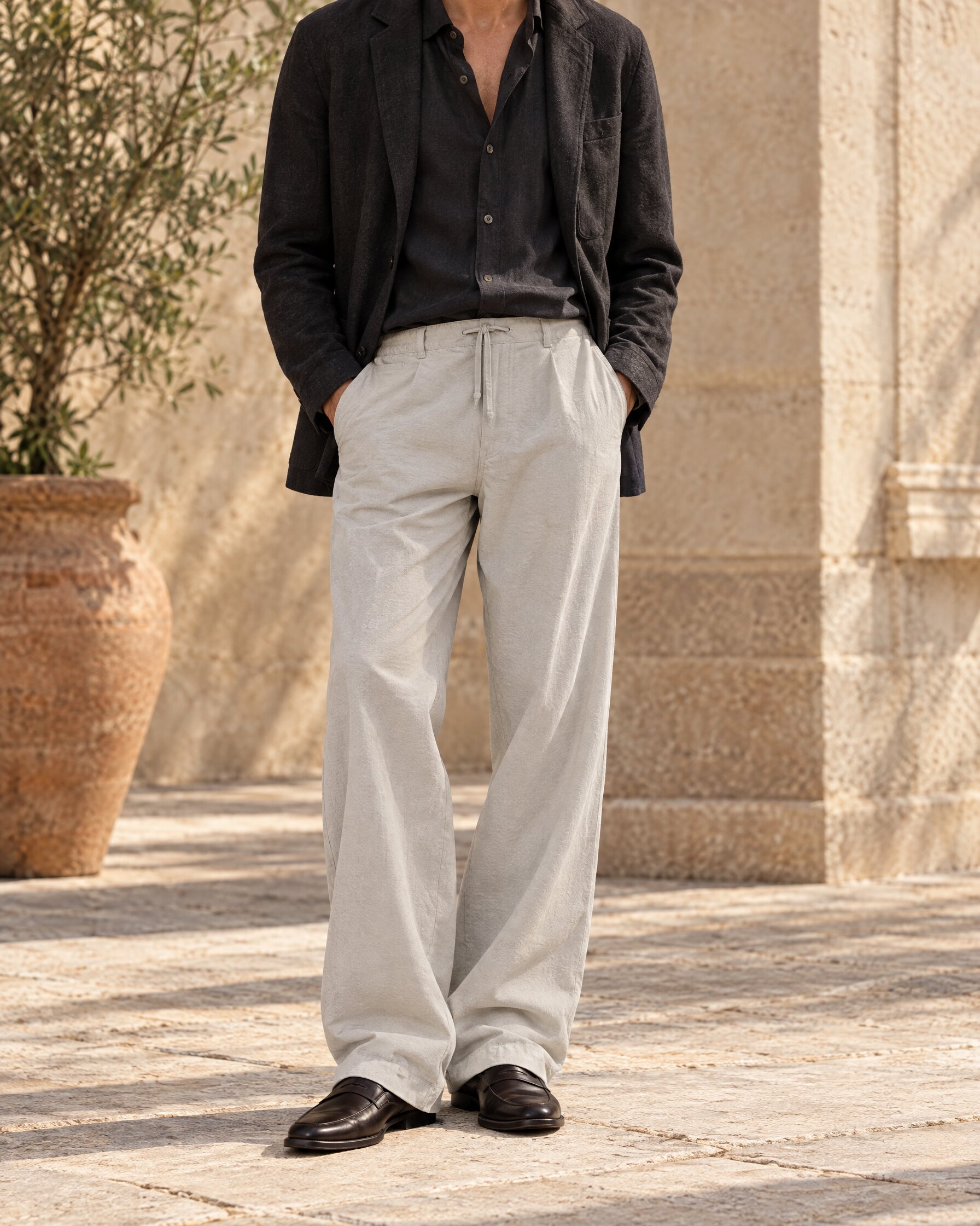 Man in pale grey drawstring trousers, dark shirt and dark jacket standing in a sunlit courtyard