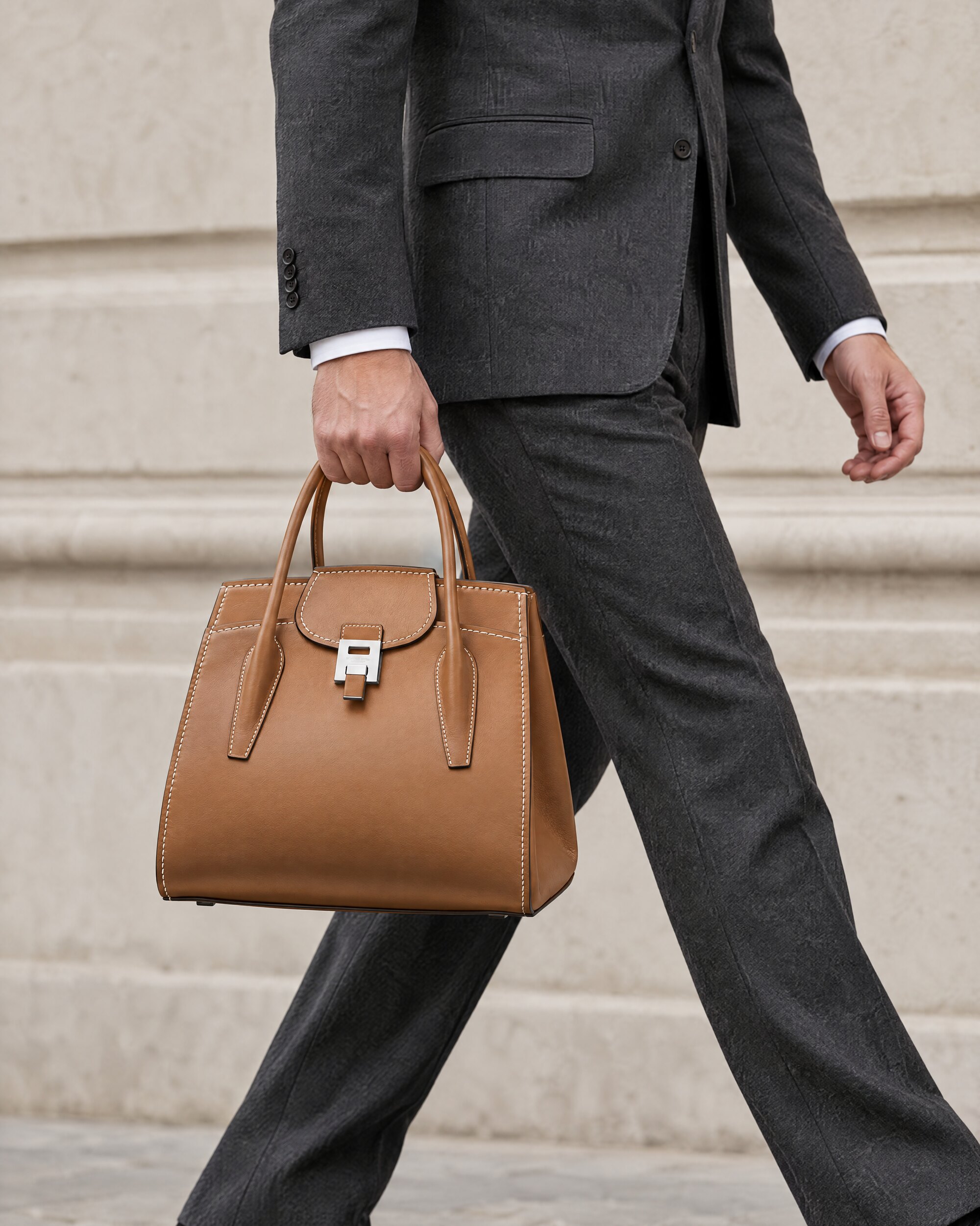 Man in a charcoal suit carrying the Michael Kors Bond Bancroft satchel by its handles