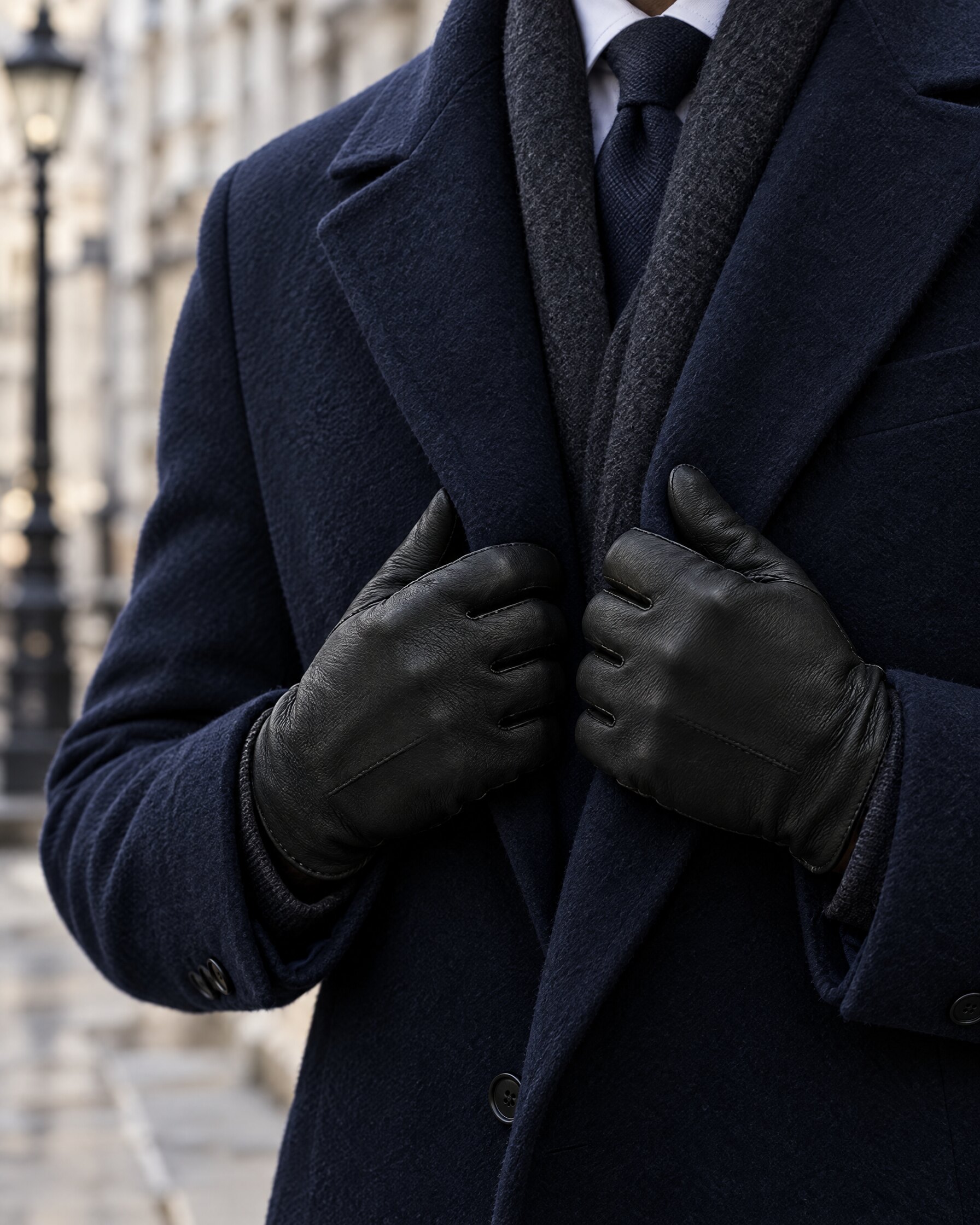 Hands in black deerskin gloves holding the lapels of a navy overcoat on a London street