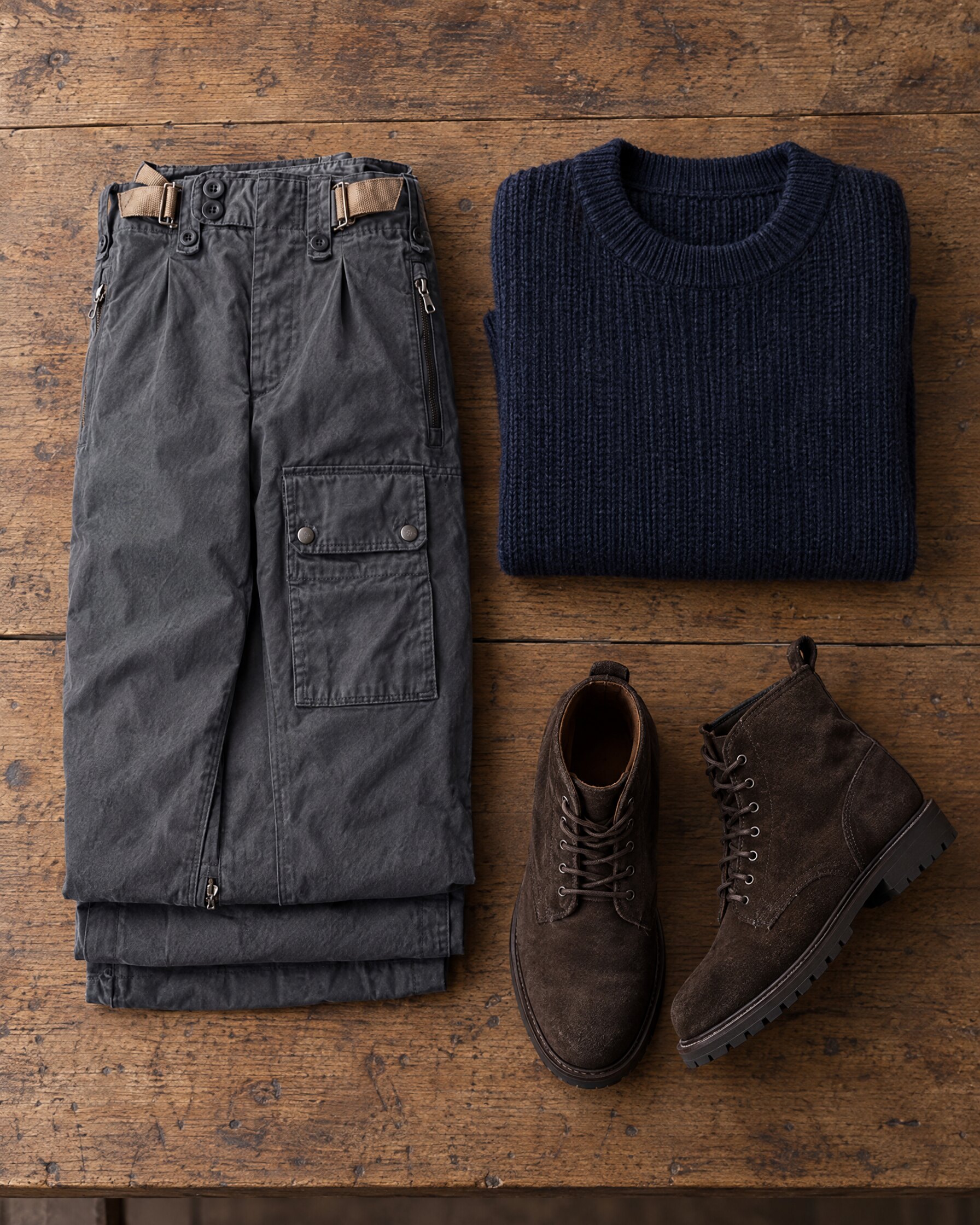 Grey N.Peal 007 combat trousers folded on an oak table beside a navy ribbed sweater and suede boots