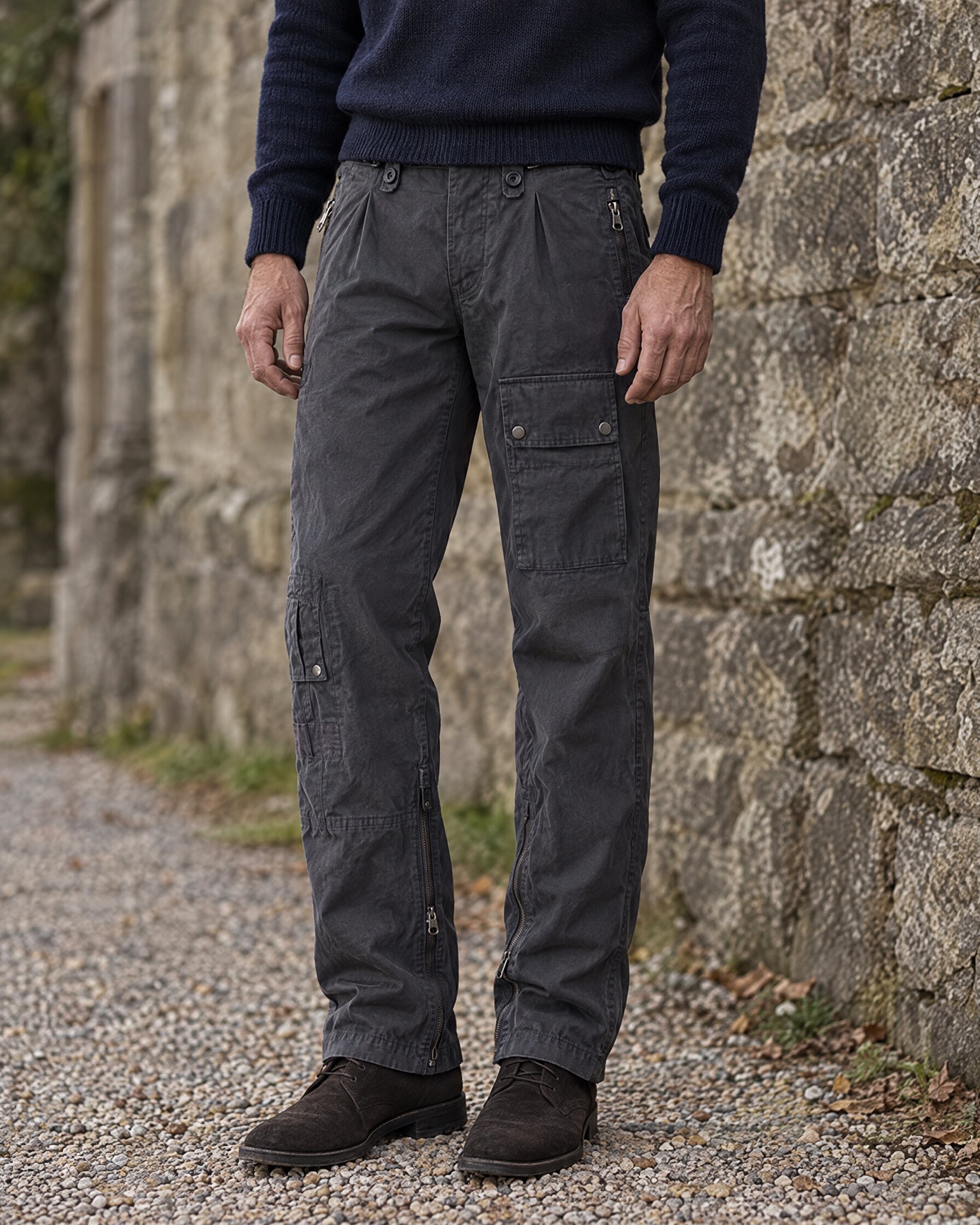 Man in grey combat trousers, navy knitwear and suede boots standing by a stone wall
