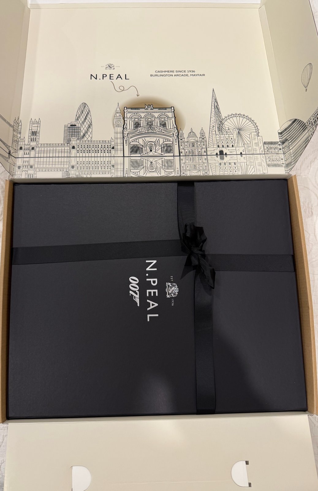 N.Peal 007 packaging for the No Time To Die Ribbed Army Sweater: cream outer box with a London skyline print open over a dark 007 gift box tied with black ribbon