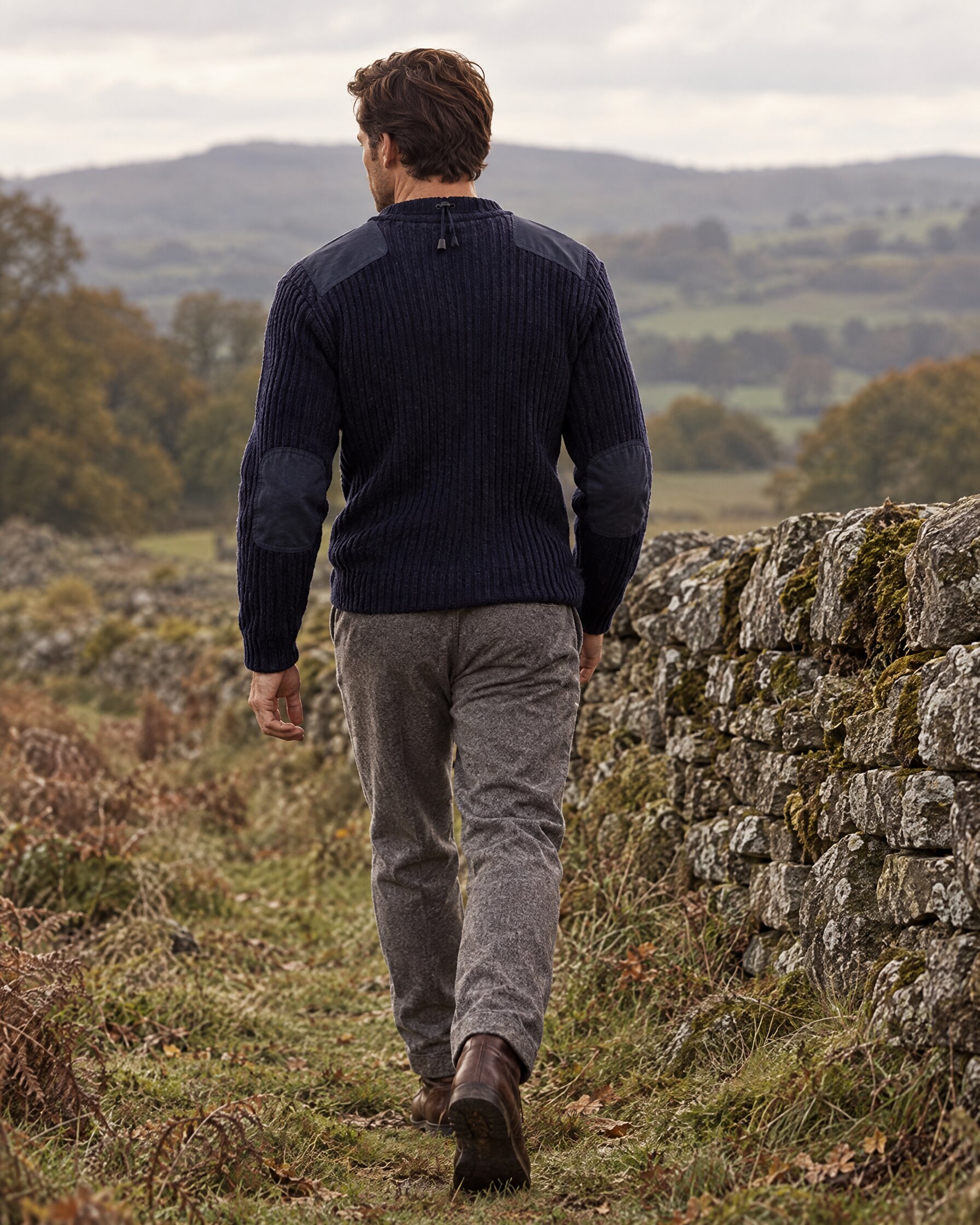 Man in the N.Peal 007 Ribbed Army Sweater in navy with grey trousers and brown boots, walking beside a stone wall