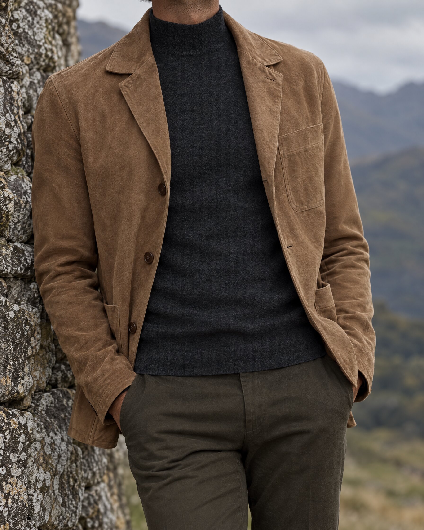 Man wearing a charcoal fine gauge mock turtle neck sweater under a tan suede jacket by a stone wall