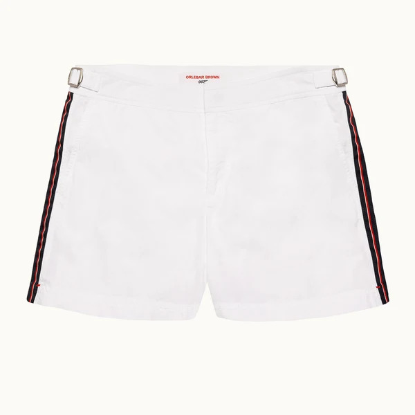 White swim shorts with taped side stripe flat lay beside sandals and a navy polo