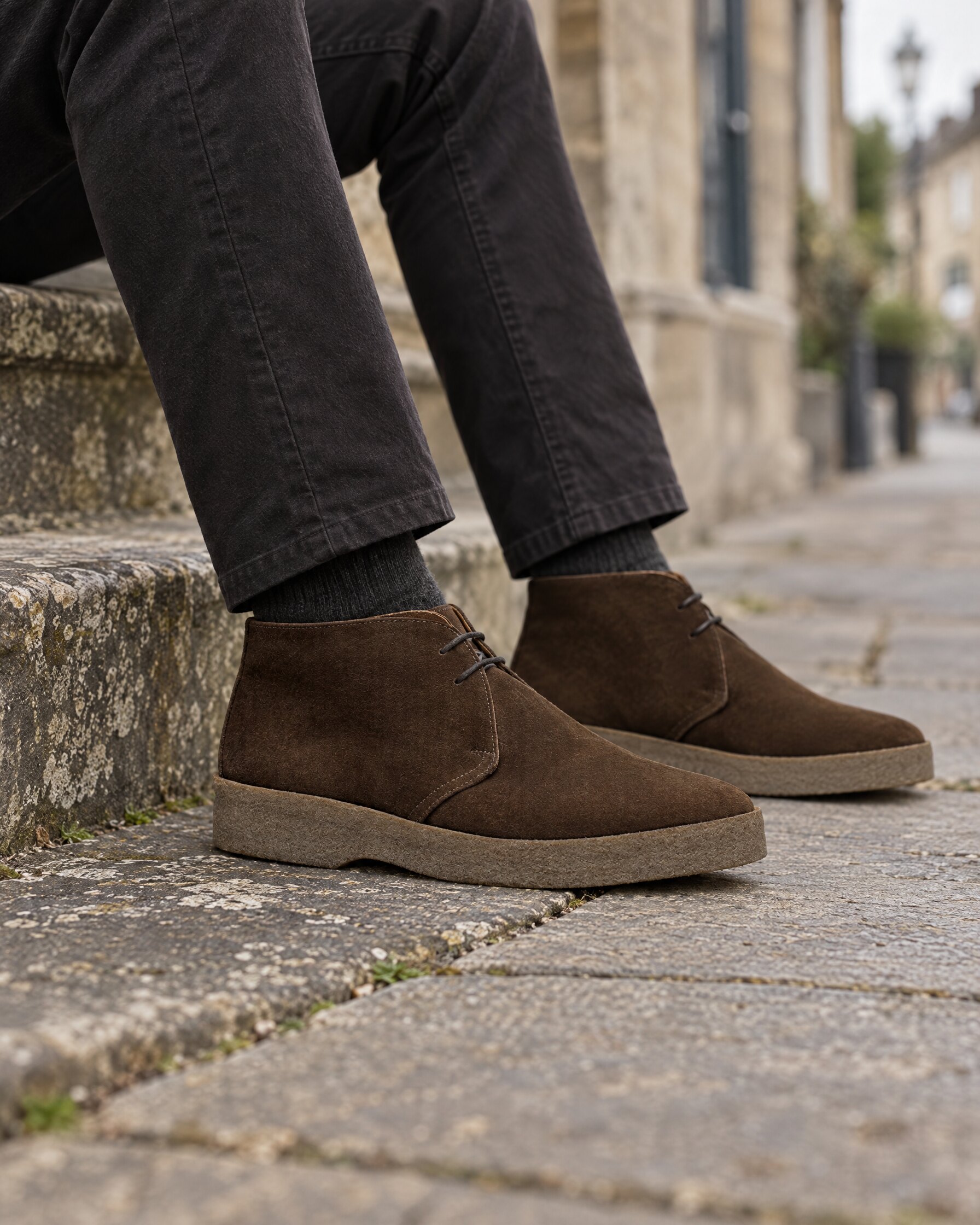 Man's legs in dark trousers and brown suede chukka boots seated on a stone step