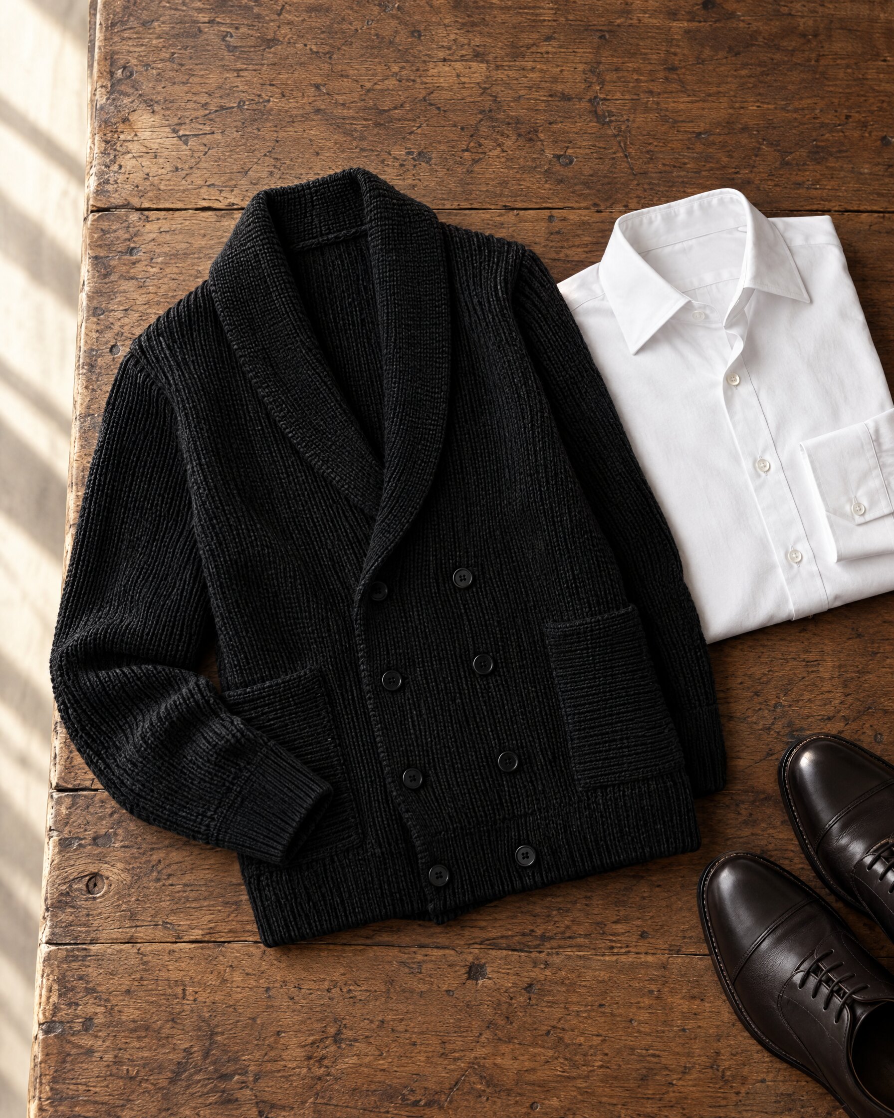 Black shawl-collar cardigan folded beside a white dress shirt on an oak surface